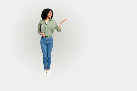 A poised African-American woman holds a laptop, gesturing invitingly towards an empty space on a white background, perfect for educational contentの写真素材