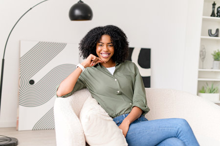 A radiant African-American woman with a charming smile sits casually on a sofa, creating an atmosphere of relaxed professionalism in a stylish living spaceの写真素材