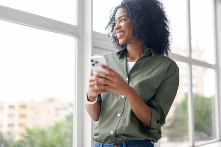 An upbeat African-American woman with a radiant smile looks away holding her smartphone, suggesting a pleasant digital interaction, with a bright window suggesting an open and comfortable living spaceの写真素材