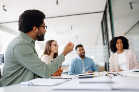 Confident businessman in casual attire sitting at a desk with a smile. Modern office setting with a productive team in the background discussing projects and ideas.の写真素材