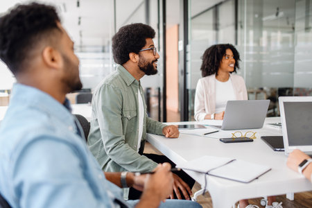 A diverse business team listens attentively during a presentation in a contemporary office. A man in a green jacket engages in discussion, while a woman with curly hair and a laptop smilesの写真素材
