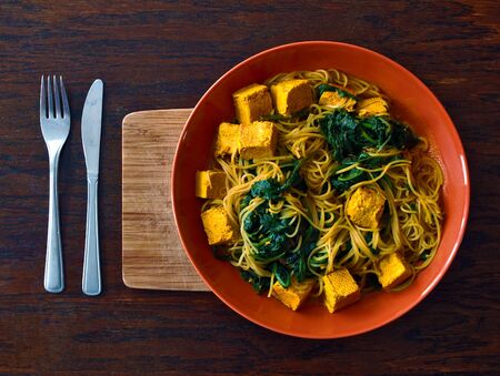 Vegetarian or vegan Indian food with noodles, spinach and tofu in the orange plate with wooden cutting board and cutlery. Suitable as a photo for a vegan or vegetarian restaurant.の写真素材