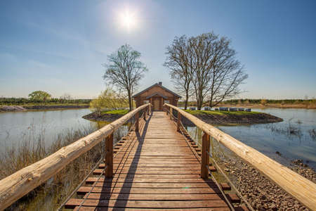 June 2020, Kosice, Czech republic - In the middle of the lake there is a house in a small grove, a wooden bridge with railings leads to it. The water is calm, the sky is blue and clear.のeditorial素材