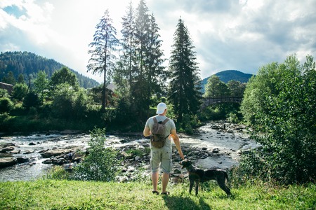young strong man stays near mountains river with dog and looking for the viewの写真素材