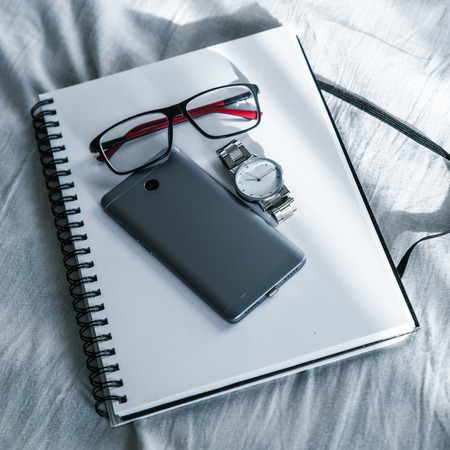 glasses, notebook, watch and a smartphone on the bed