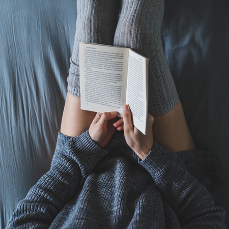 woman read book in bed with gray sheets. overhead view