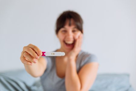 young happy woman sitting on bed looking on positive pregnancy test. blurred backgroundの写真素材