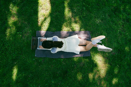 young pretty woman laying on blanket on green grass with laptop. overhead view