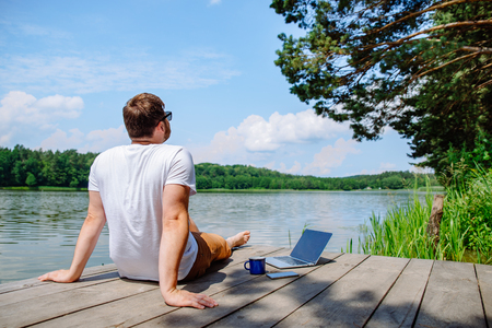 man working on laptop while sitting on wooden dock. legs in river waterの写真素材