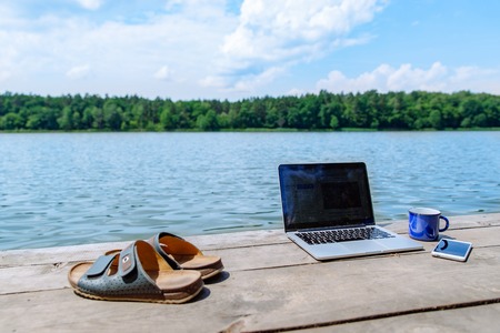 laptop with cup and phone one wooden dock. river on background. summer time. work with youの写真素材