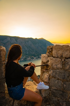 young adult woman sitting on top of hill on sunset with beautiful view of kotor.の写真素材