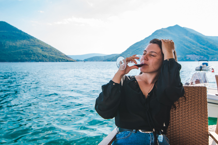 smiling young woman drinking red wine at seaside with beautiful mountains view. resting in cafeの写真素材