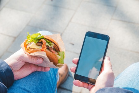 man holding burger and mobile phone. white screen. copy spaceの写真素材