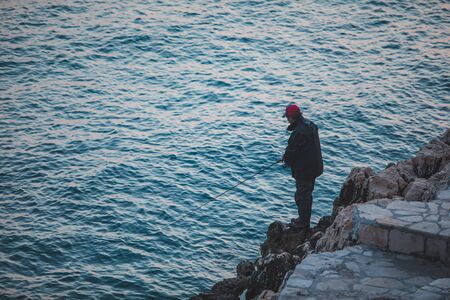 ROVINJ, CROATIA - May 22, 2019: man sitting on cliff and fishing in sea. backgroundのeditorial素材