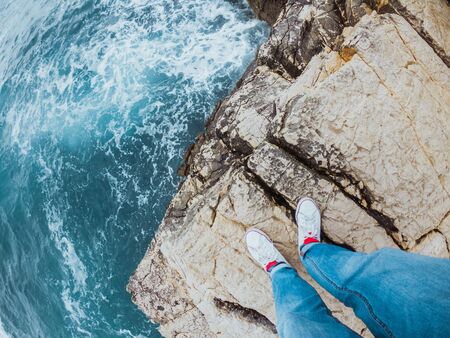 man legs in jeans and white sneakers at the edge of the cliff view of blue stormy sea