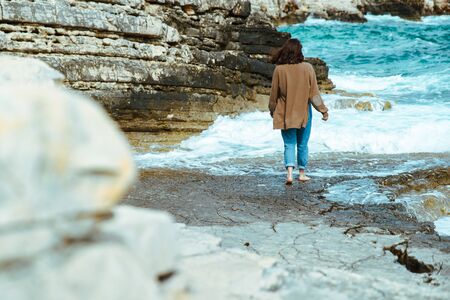 woman walking by rocky sea beach at sunny windy day. summer vacation. carefree concept . waves on backgroundの写真素材