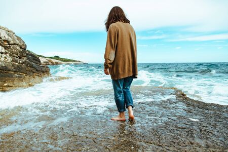 woman walking by rocky sea beach at sunny windy day. summer vacation. carefree concept . waves on backgroundの写真素材