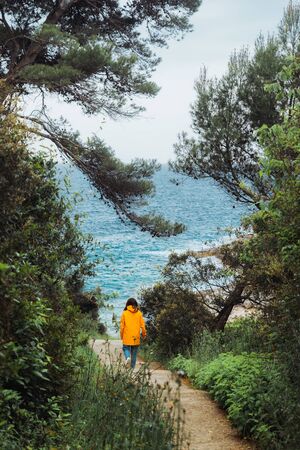 woman in yellow raincoat walking to the sea by trail between trees. summer vacationの写真素材