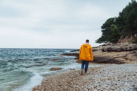 man walking by rocky beach in yellow raincoat. overcast windy weather. seasideの写真素材