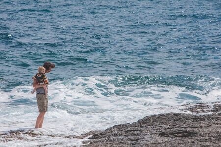 PULA, CROATIA - May 20, 2019: young father holding kid on hands showing waves in sea. summer vacationのeditorial素材