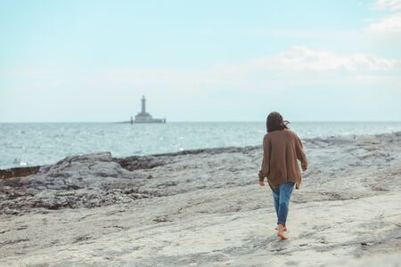 woman walking by rocky sea beach in wet jeans lighthouse on background. windy weather. summer vacation. carefree conceptの写真素材