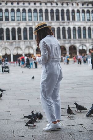 woman in white clothes with straw hat having fun with pigeons at venice city square piazza san marco