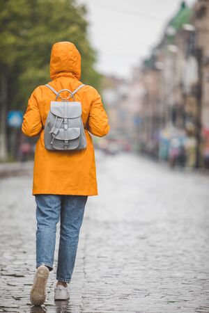 woman in yellow raincoat walking by city streets under rain. view from backの写真素材