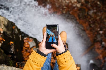woman taking picture on phone of her legs with waterfall on background autumn seasonの写真素材