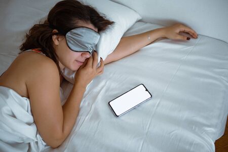 woman sleeping in sleep mask smartphone near her morning alarm white screen copy spaceの写真素材