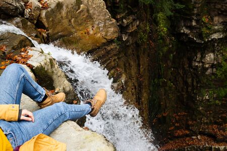 woman in blue jeans and brown boots sitting on the edge looking at waterfall autumn time fall season
