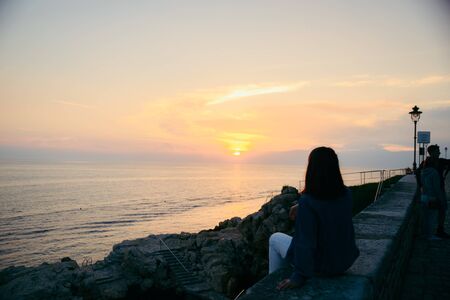 young pretty woman sitting at cliff looking on beautiful sunset. summer travel concept