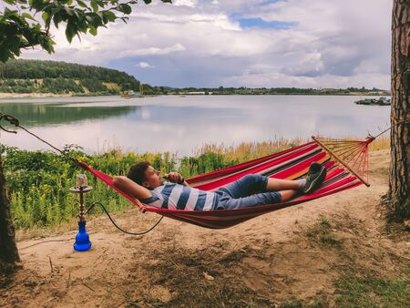 man laying at hammock smoking hookah looking at lake beach summer time