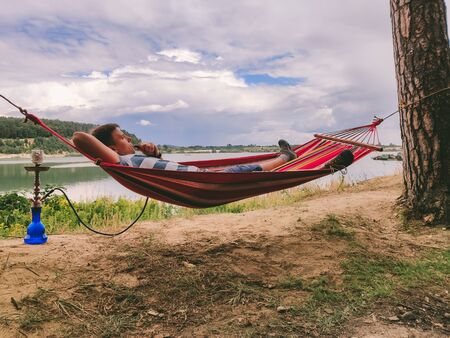 man laying at hammock smoking hookah looking at lake beach summer time