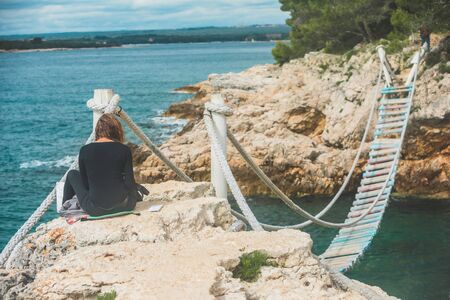 woman sitting at the edge drawing picture of seascape suspension bridge