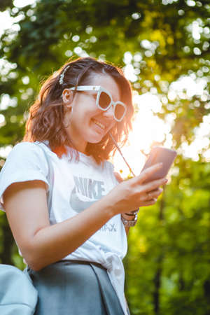 Lviv, Ukraine - July 30, 2019: young stylish woman walking by public park on sunset using phone drinking cool smoothie. lifestyle conceptのeditorial素材