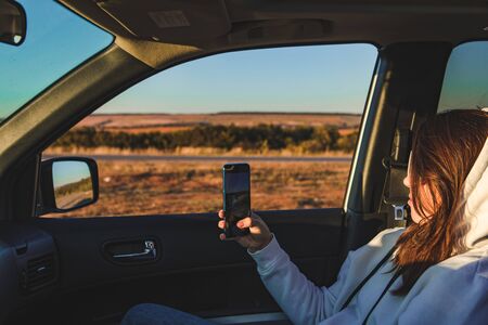 car travel concept woman taking picture on cellphone from car evening sunset timeの写真素材