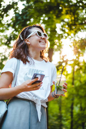 Lviv, Ukraine - July 30, 2019: young stylish woman walking by public park on sunset using phone drinking cool smoothie. lifestyle conceptのeditorial素材