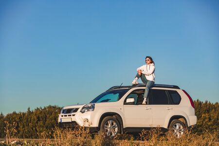 stylish woman in casual outfit sitting on the roof of the car at sunset road trip conceptの写真素材