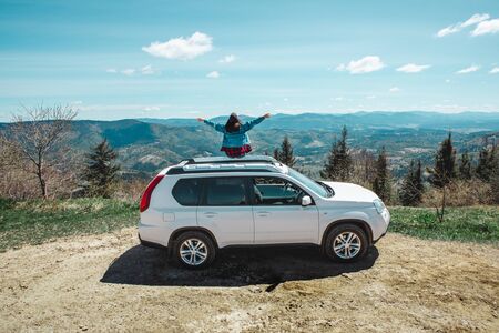 young woman sitting on the top of the suv car at mountain peak enjoying the landscape view at summer sunny dayの写真素材