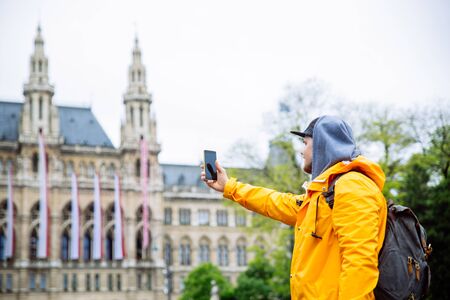 tourist traveler man taking picture on phone vienna city hall on background spring rainy weather yellow raincoatの写真素材