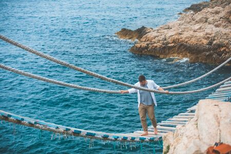 man walking by suspension bridge cross sea bay rocky cliff summer timeの写真素材