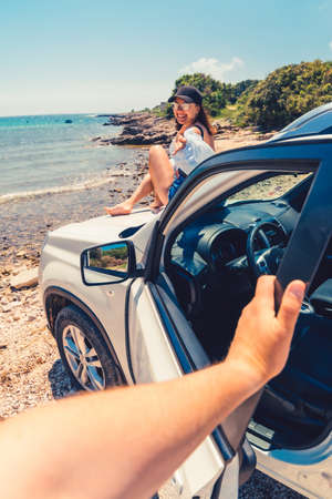 first person point of view man closing door of the car woman sitting on the hood view on summer beach car travel conceptの写真素材