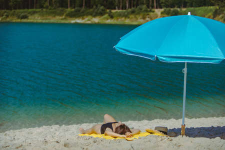 woman laying on yellow blanket at sand beach with blue sun umbrella tanningの写真素材