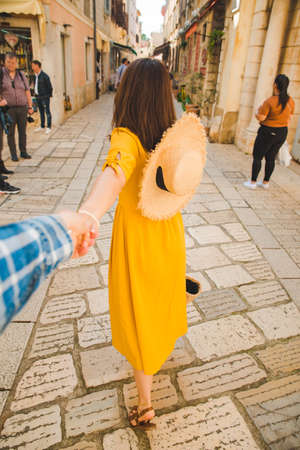follow me concept. woman in yellow sundress in straw hat walking forward by small resort city street holding man hand. summer vacationの写真素材