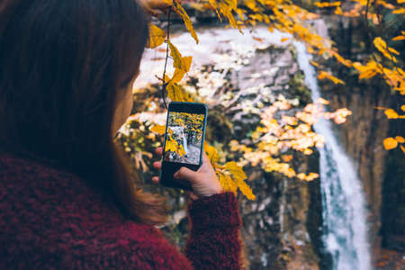 woman taking picture of autumn waterfall on the phone hiking concept