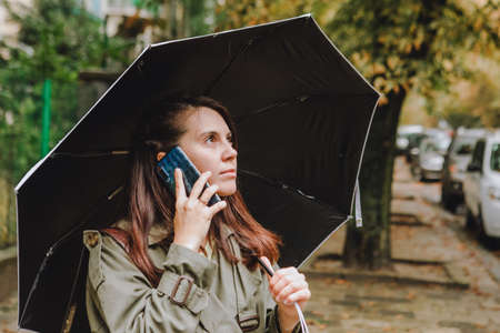young pretty woman talking on the phone at city street hiding from rain under umbrellaの写真素材