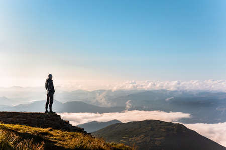 man standing at the edge of the cliff looking at mountains. clouds belowの写真素材