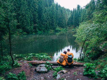 couple hikers in yellow raincoat looking at mountain lake backpackersの写真素材