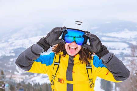 young pretty smiling woman in ski outfit with goggles and helmet mountains on background stick tongue outの写真素材