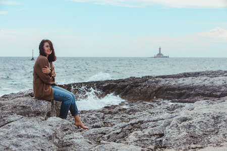 woman sitting by rocky sea beach in wet jeans lighthouse on background